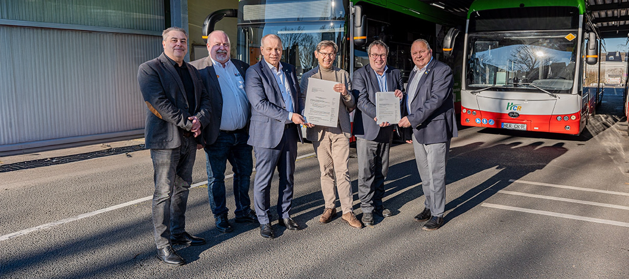 Six men standing in front of a bus depot holding a grant certificate, flanked by two HCR buses