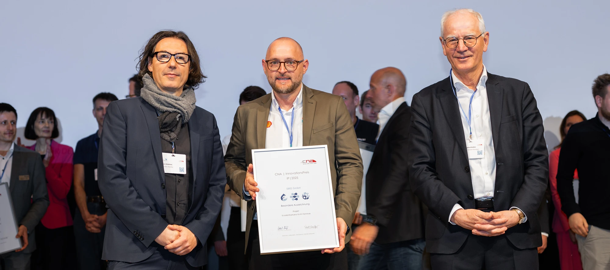 Three people standing on stage holding a framed certificate, with several other people in the background at an award ceremony