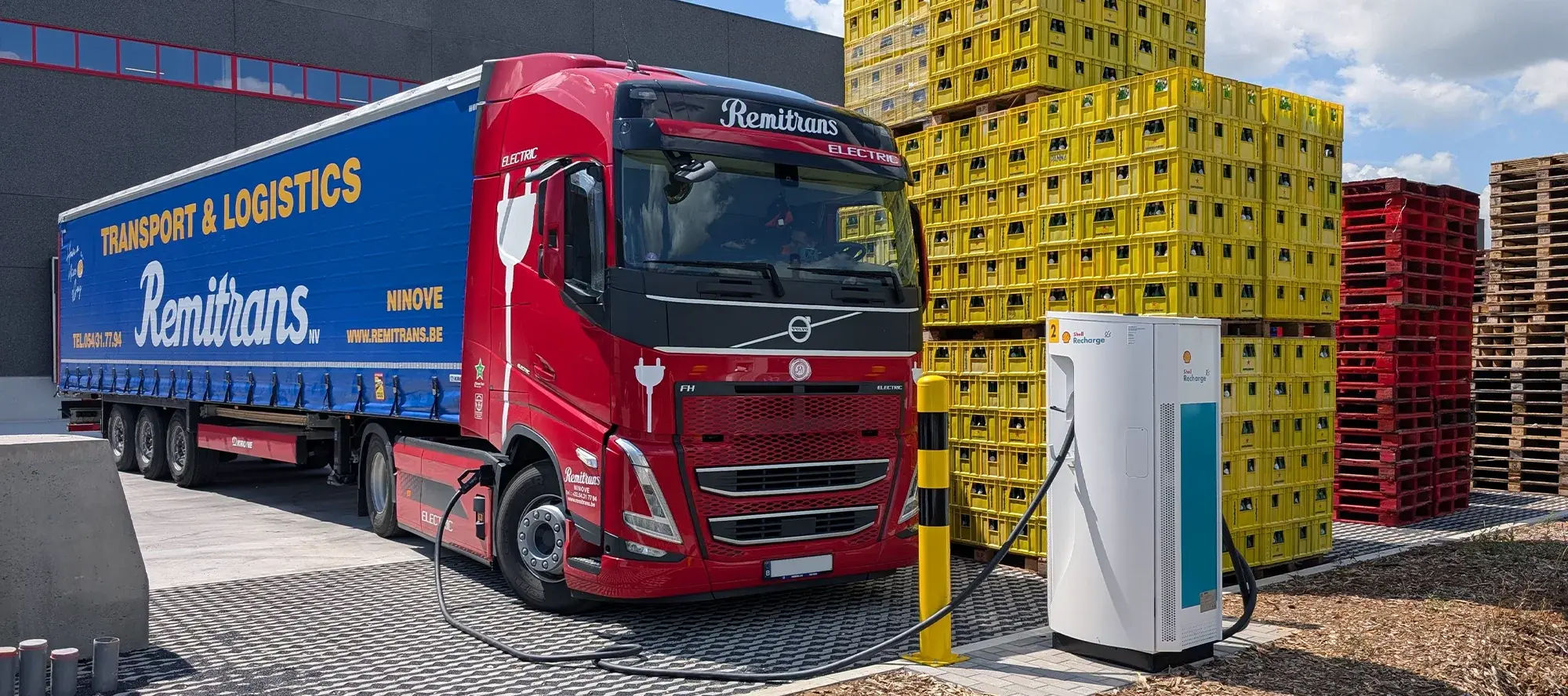 Electric Remitrans truck charging at a Shell Recharge station at the semi-public depot in Ath, Belgium, surrounded by stacked crates and pallets.