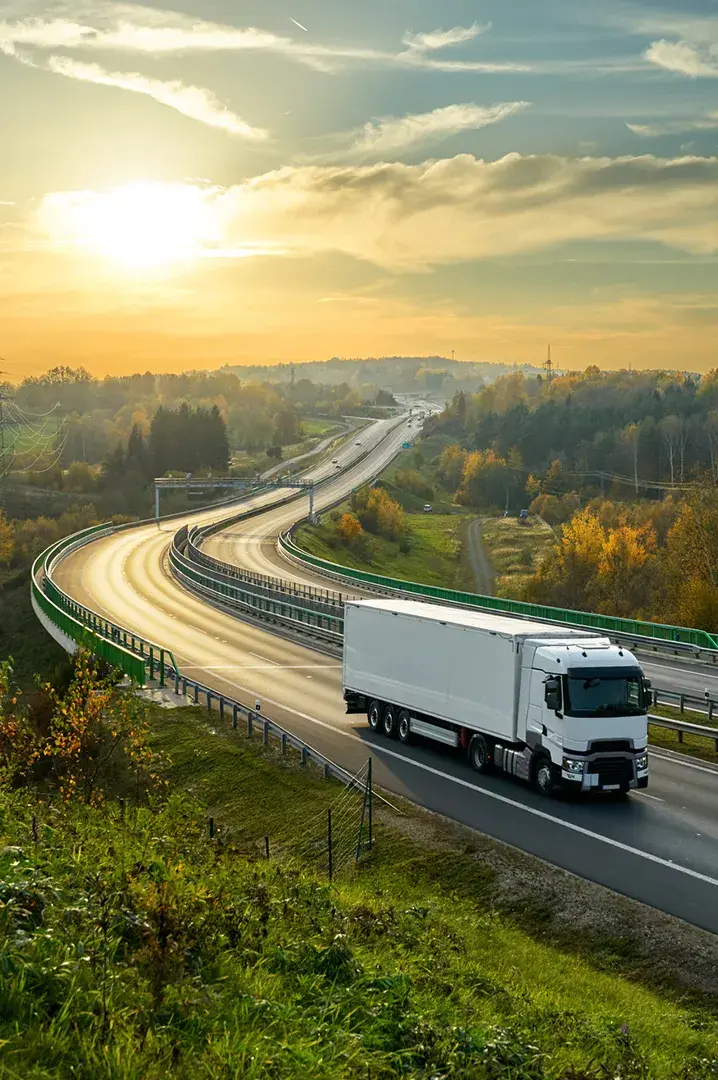 Elektro-Lkw fährt bei Sonnenaufgang auf einer kurvigen Landstraße durch eine ländliche Gegend.